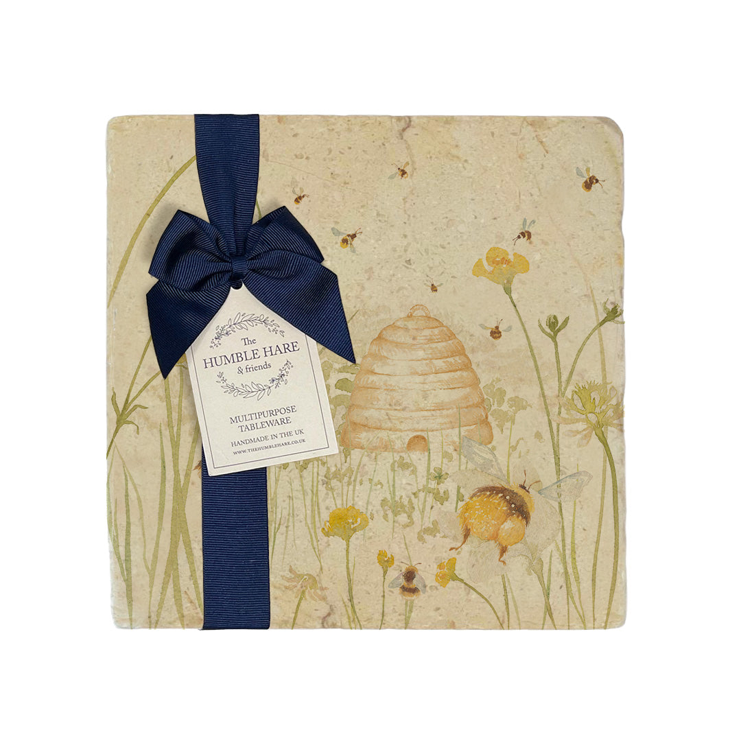 A multipurpose marble platter with a watercolour design featuring bees and a beehive in a buttercup meadow, packaged with a luxurious dark blue bow and branded gift tag.