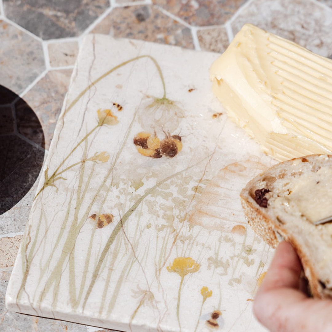 A medium square marble platter being used as a butter dish, the platter features a watercolour design of bees and a beehive in a buttercup meadow.