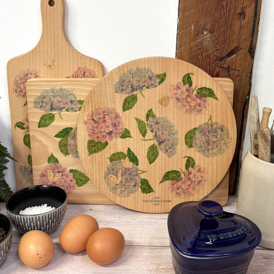 Round wooden chopping board on a kitchen worktop. The beech wood board features a watercolour floral pattern of blue and pink hydrangea flowers and butterflies.