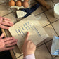 A grandma and granddaughter pointing at a handwritten recipe marble platter on a tiled kitchen countertop with eggs, flour and other baking items.