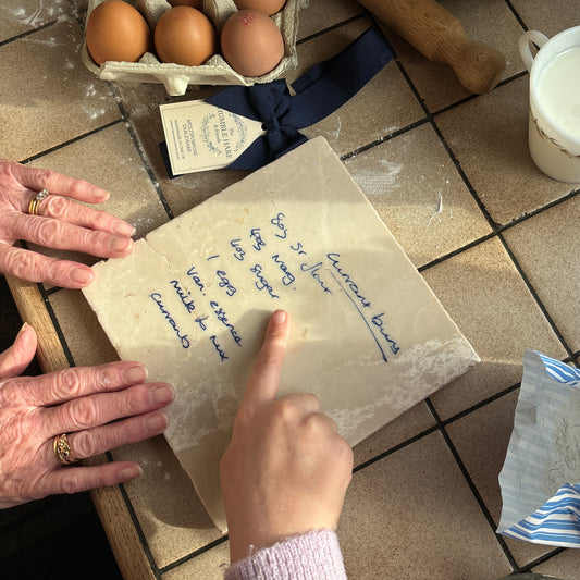 A grandma and granddaughter pointing at a handwritten recipe marble platter on a tiled kitchen countertop with eggs, flour and other baking items.