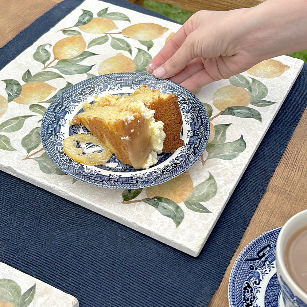 A large marble placemat with a pretty lemon pattern. A hand is placing a plate with a slice of cake on the placemat.