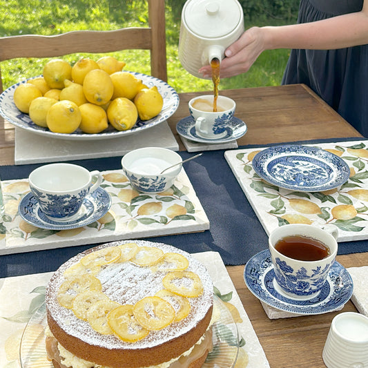 A table in the garden set with cake, teacups and saucers for afternoon tea, all placed on multipurpose marble platters featuring a watercolour lemon pattern.