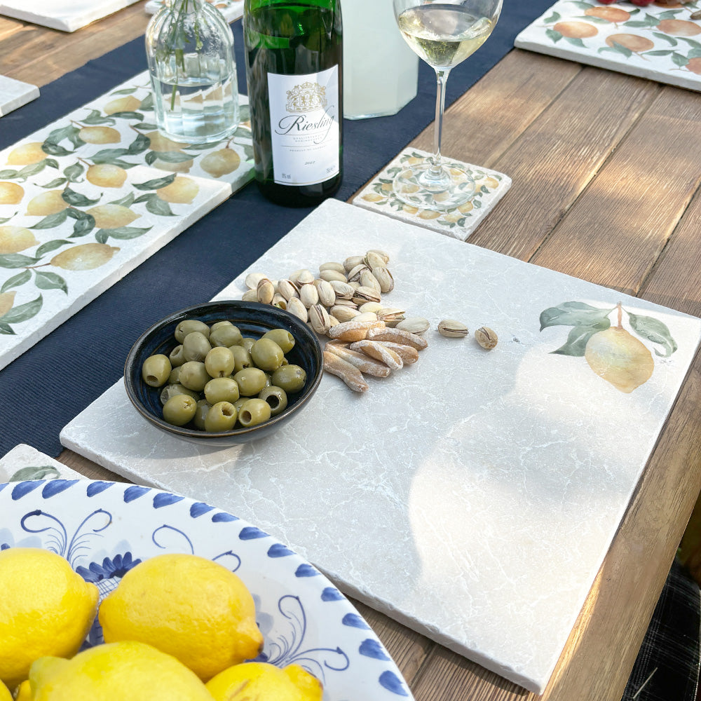 A large marble cheeseboard with a minimalistic lemon design, it is being used to serve nuts, olives and nibbles on a garden table.