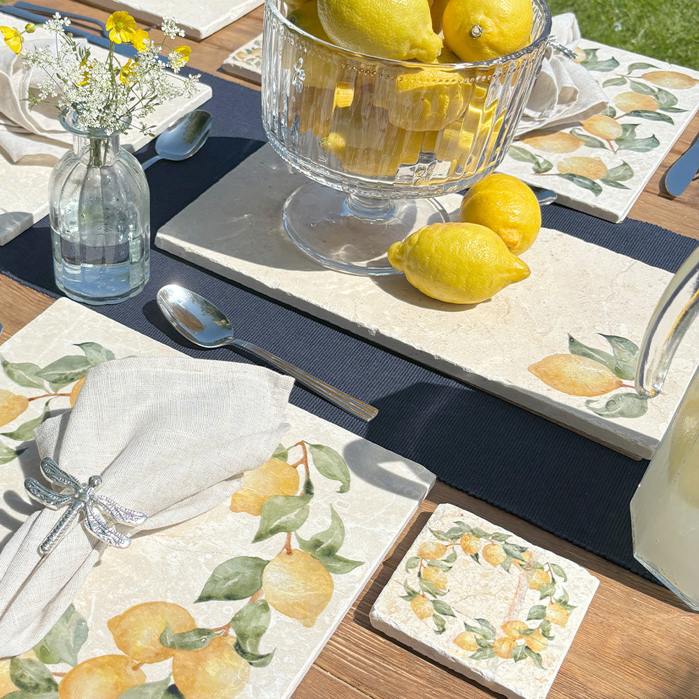 A garden table set with marble placemats and serving platters featuring a minimalistic lemon watercolour design.