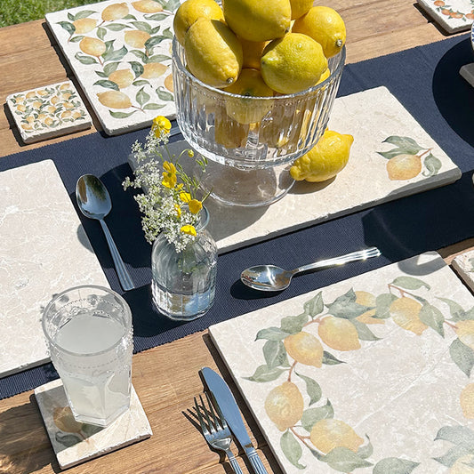 A garden table set with marble placemats and serving platters featuring a minimalistic lemon watercolour design.