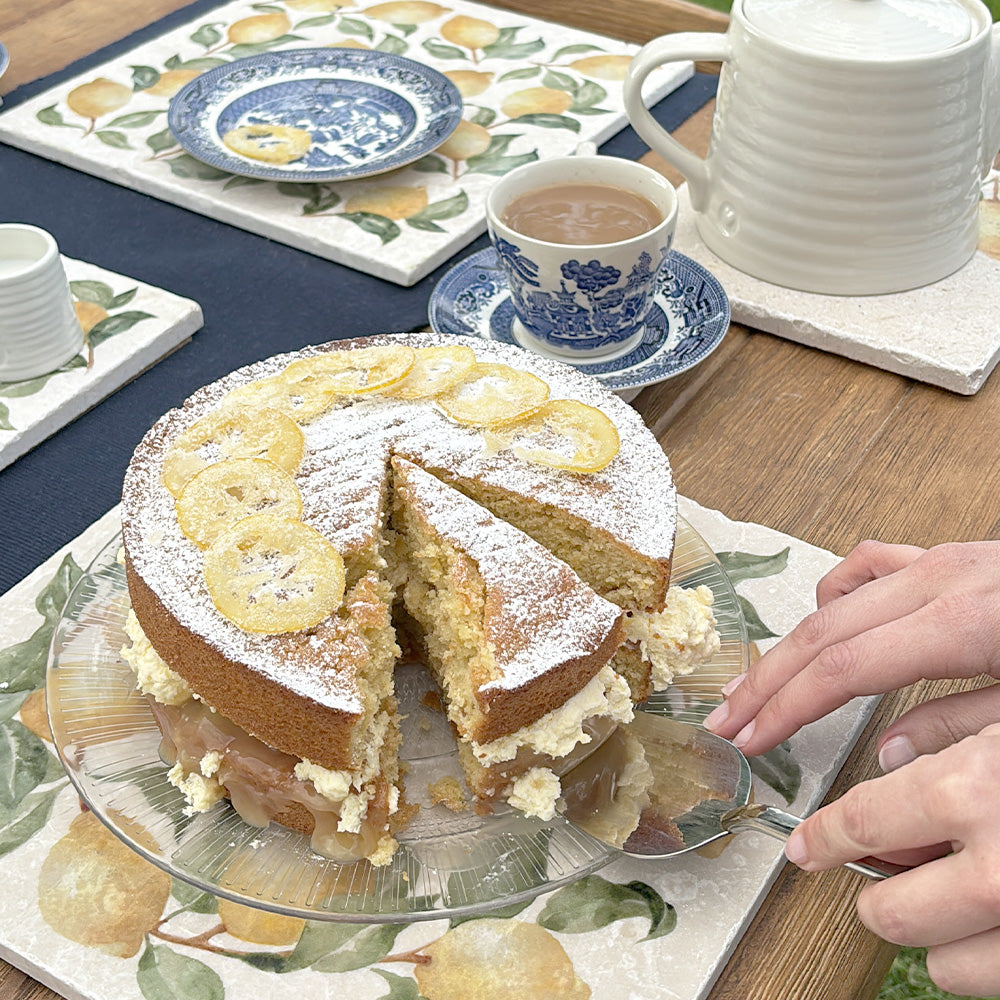 A large marble platter featuring a watercolour lemon wreath design. The platter is being used as a serving board for a lemon sponge cake.