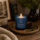 A blue glass jar candle with a lit flame sits on a wooden tray on a rustic table, styled with a pinecone, evergreen sprigs, a mug, a framed print, and a knitted blanket. The candle label shows that the scent is 'Wild Woodland' a woody, earthy scent.