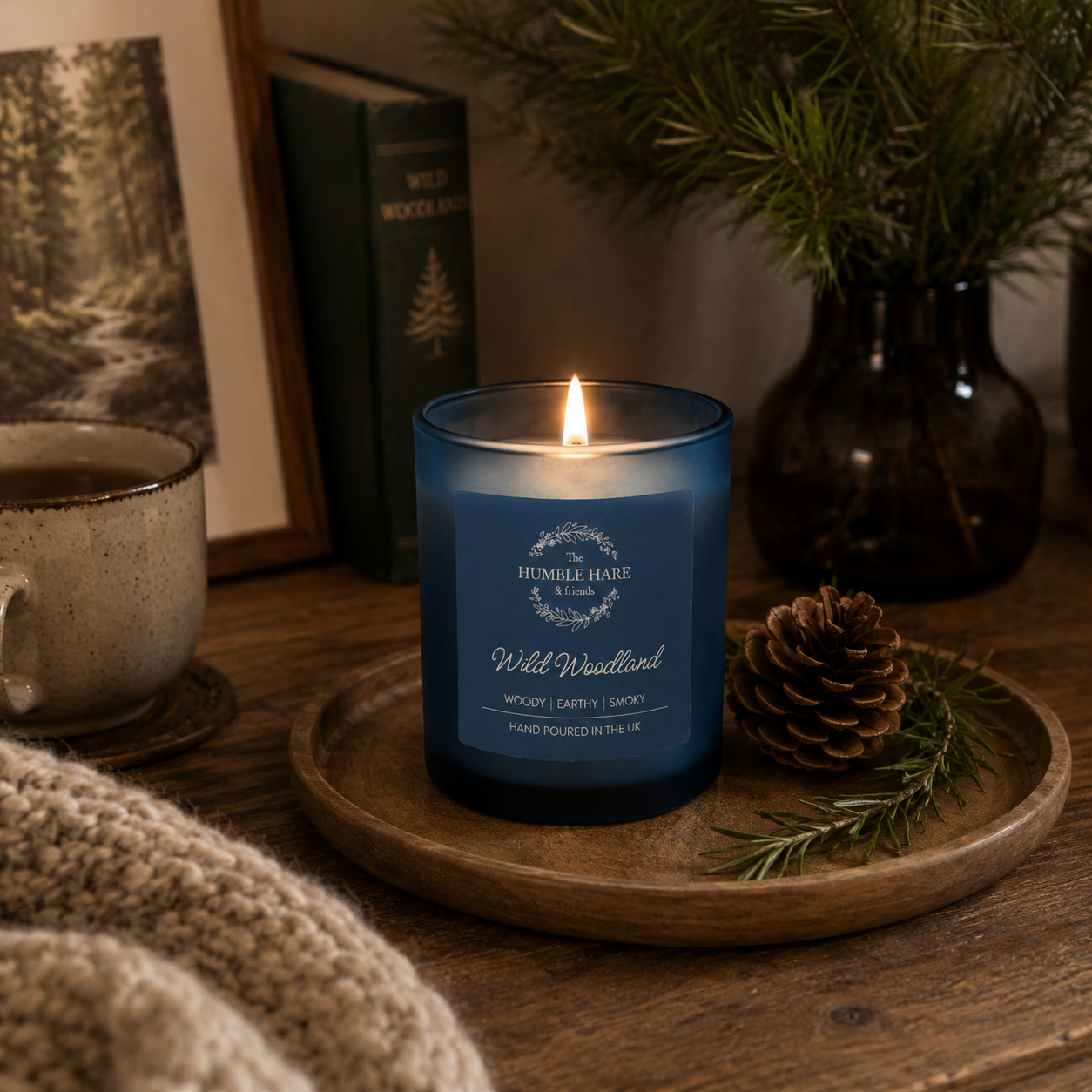 A blue glass jar candle with a lit flame sits on a wooden tray on a rustic table, styled with a pinecone, evergreen sprigs, a mug, a framed print, and a knitted blanket. The candle label shows that the scent is 'Wild Woodland' a woody, earthy scent.