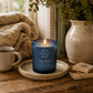 A blue glass jar candle with a lit flame sits on a small round tray on a wooden table, styled with a mug, books, a knitted blanket, and a ceramic vase in a cozy indoor setting.