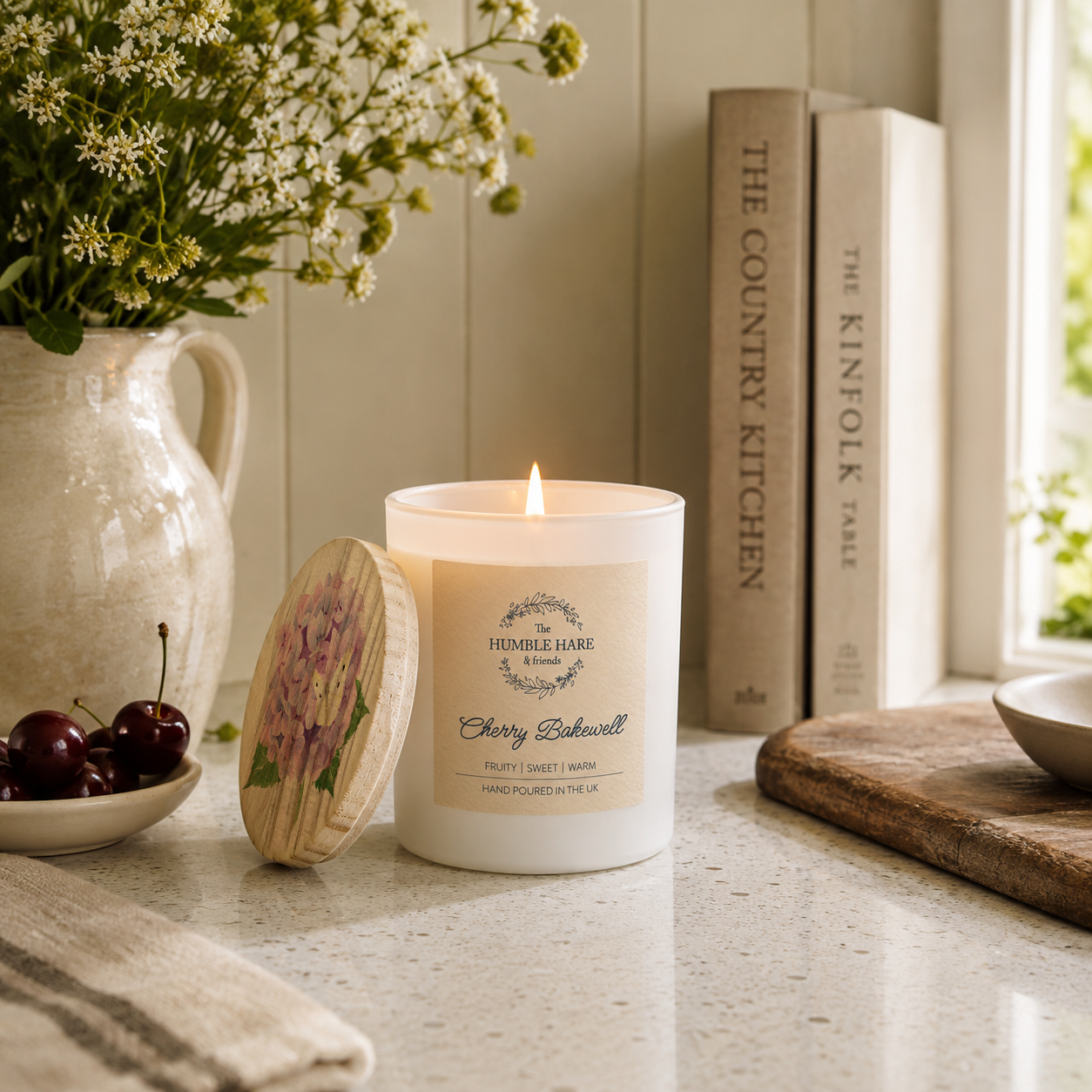 A lit white candle with a cream label with a Cherry Bakewell scent sits on a kitchen counter beside its wooden lid, with flowers in a vase, books, cherries, and a cutting board nearby.