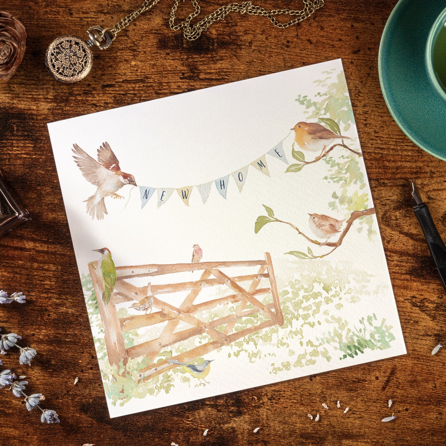 A greetings card laid flat on a table surrounded by writing items including a fountain pen and ink. The card reads New Home on bunting held by a sparrow and a robin above an open garden gate in a watercolour style.