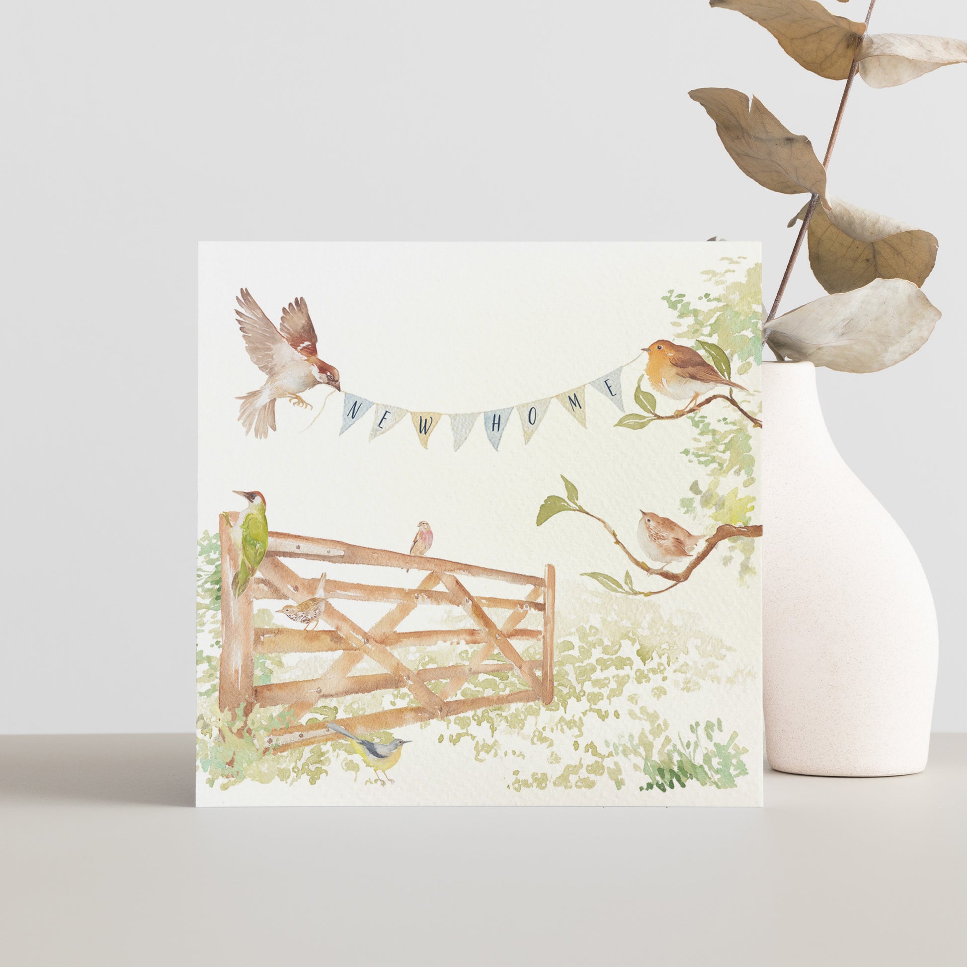 A greetings card reading New Home on bunting held by a sparrow and a robin above an open garden gate in a watercolour style.