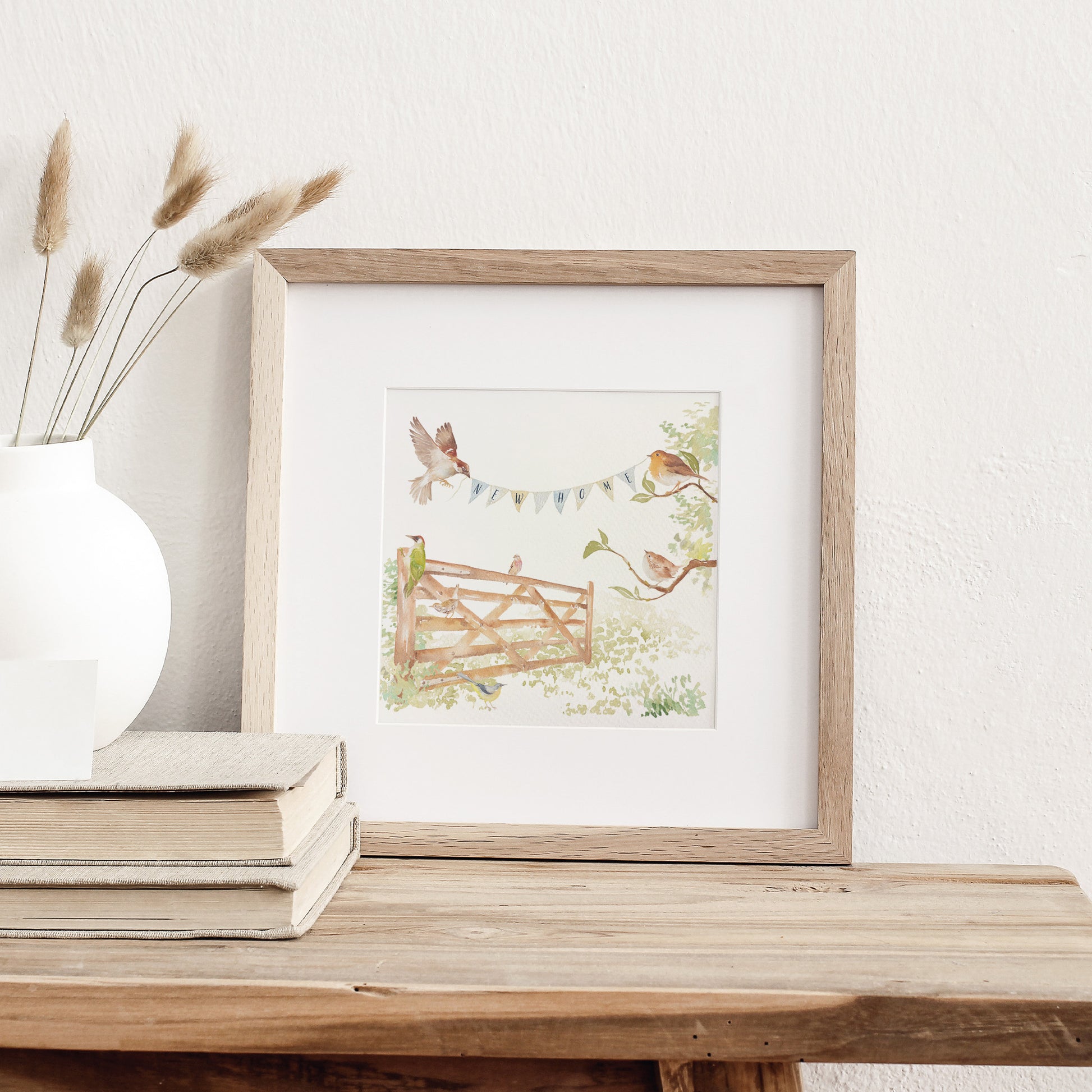 A greetings card displayed as an art print in a neutral coloured frame propped up on a bookshelf. The card reads New Home on bunting held by a sparrow and a robin above an open garden gate in a watercolour style.