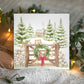 A square Christmas card featuring a winter forest scene with mice, snow-covered evergreen trees, a rustic cabin, a wooden gate, a green wreath, and a red bow. The card is displayed on a textured cream knit blanket with warm string lights and decorative greenery in the background.