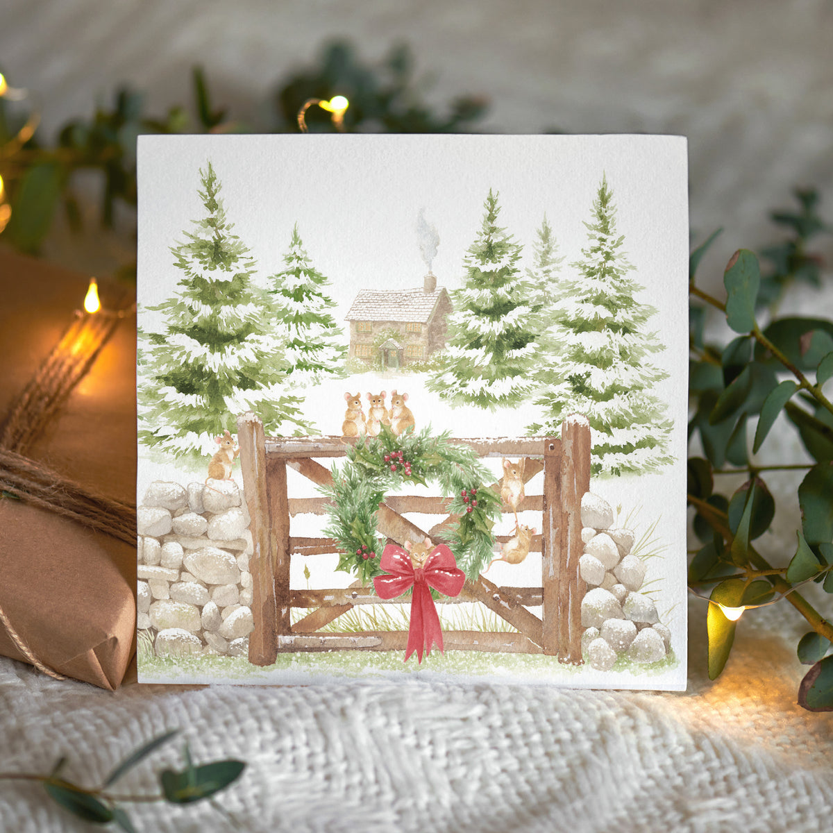A square Christmas card featuring a winter forest scene with mice, snow-covered evergreen trees, a rustic cabin, a wooden gate, a green wreath, and a red bow. The card is displayed on a textured cream knit blanket with warm string lights and decorative greenery in the background.