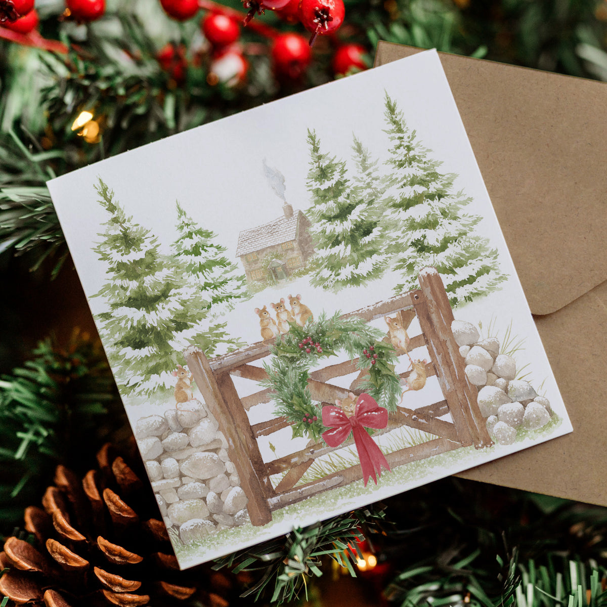 A square Christmas greeting card with an illustrated winter scene showing snow-covered evergreen trees, a small cabin, a wooden gate with mice , and a green wreath with a red bow, placed on top of a brown envelope and surrounded by holiday greenery and pinecones.