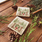Set of four square coasters featuring a rustic Christmas scene with mice playing on a wooden gate, green wreath, red bow, snowy pine trees, and a small cottage.
