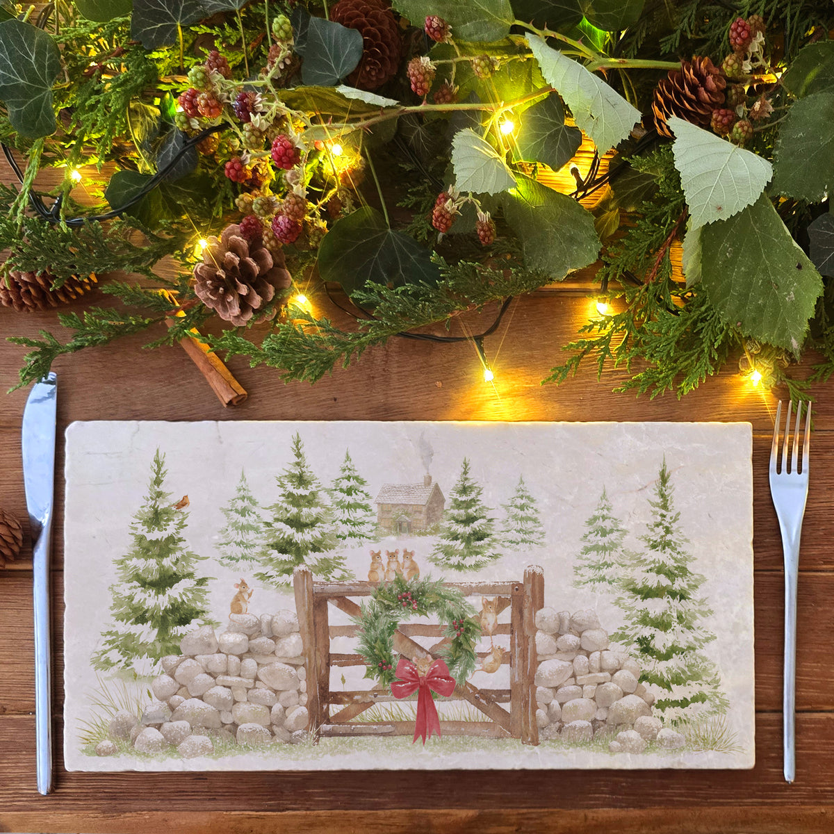 A rectangular marble sharing platter featuring a rustic winter forest scene with mice playing on a wooden gate, green wreath, red bow, stone wall, evergreen trees, and a small cabin.