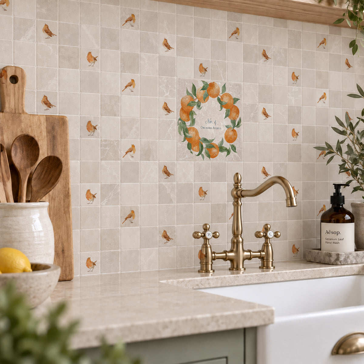 A kitchen backsplash made of beige marble tiles decorated with orange and green fruit watercolour design including a central wreath design with the text 'Our Happy Place' above a brass faucet and stone countertop.