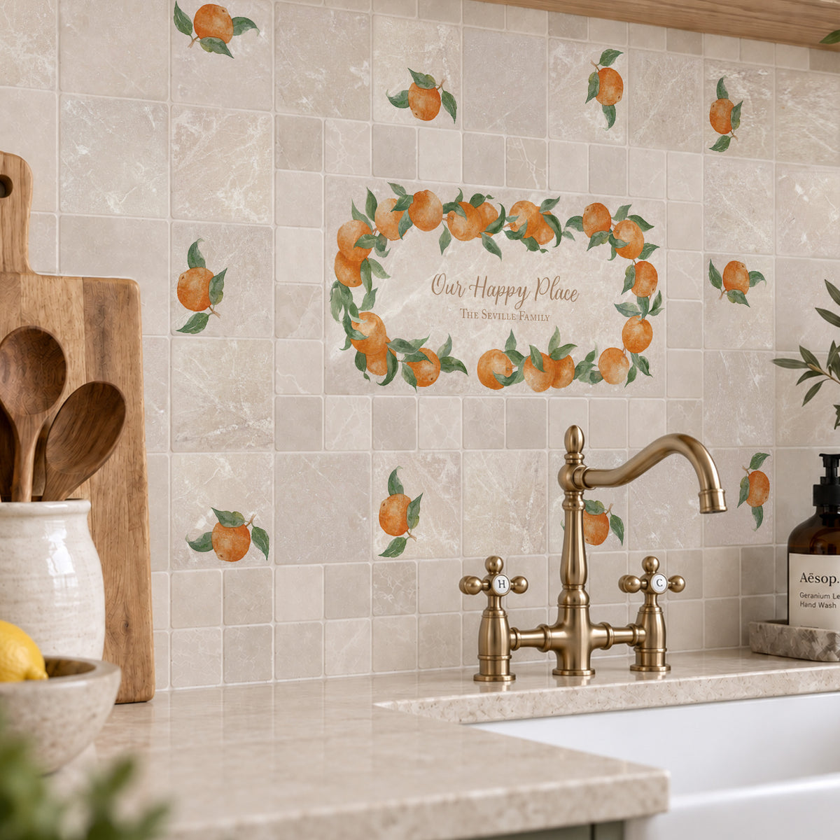 A kitchen backsplash made of beige marble tiles decorated with orange and green fruit watercolour design including a central wreath design with the text 'Our Happy Place' above a brass faucet and stone countertop.