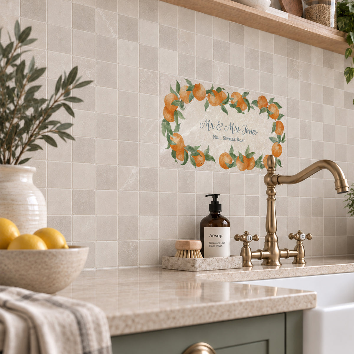 A decorative wall decal featuring an orange and green citrus wreath with the text 'Mr & Mrs Jones' and 'New Orleans Born' is shown on a beige tiled kitchen backsplash above a sink with a brass faucet.