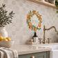 A kitchen backsplash made of beige marble tiles decorated with orange and green fruit watercolour design including a central wreath design with the text 'Our Happy Place' above a brass faucet and stone countertop.