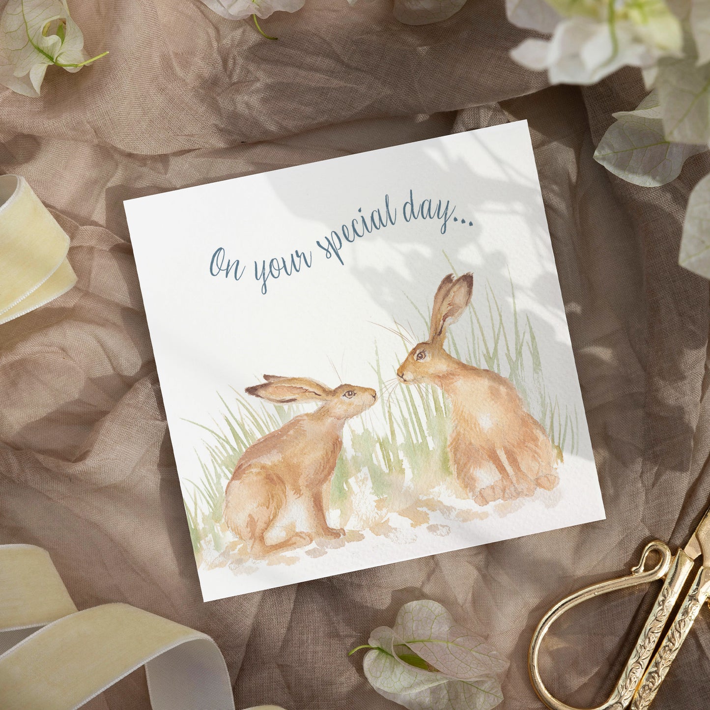 A greetings card laid flat on a table surrounded by gift wrapping items including scissors and ribbon. The card reads On Your Special Day in dark blue text above a hare couple in a watercolour style.