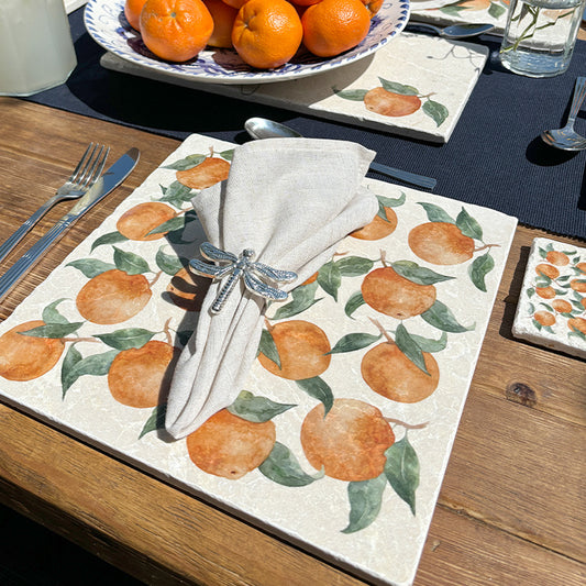 An al fresco dinging table set with marble placemats and coasters featuring a maximalist  pattern of tumbling Mediterranean oranges and leaves.