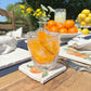 An al fresco dining table set with a beautiful cocktail glass on top of a square marble coaster featuring a minimalistic watercolour orange design.