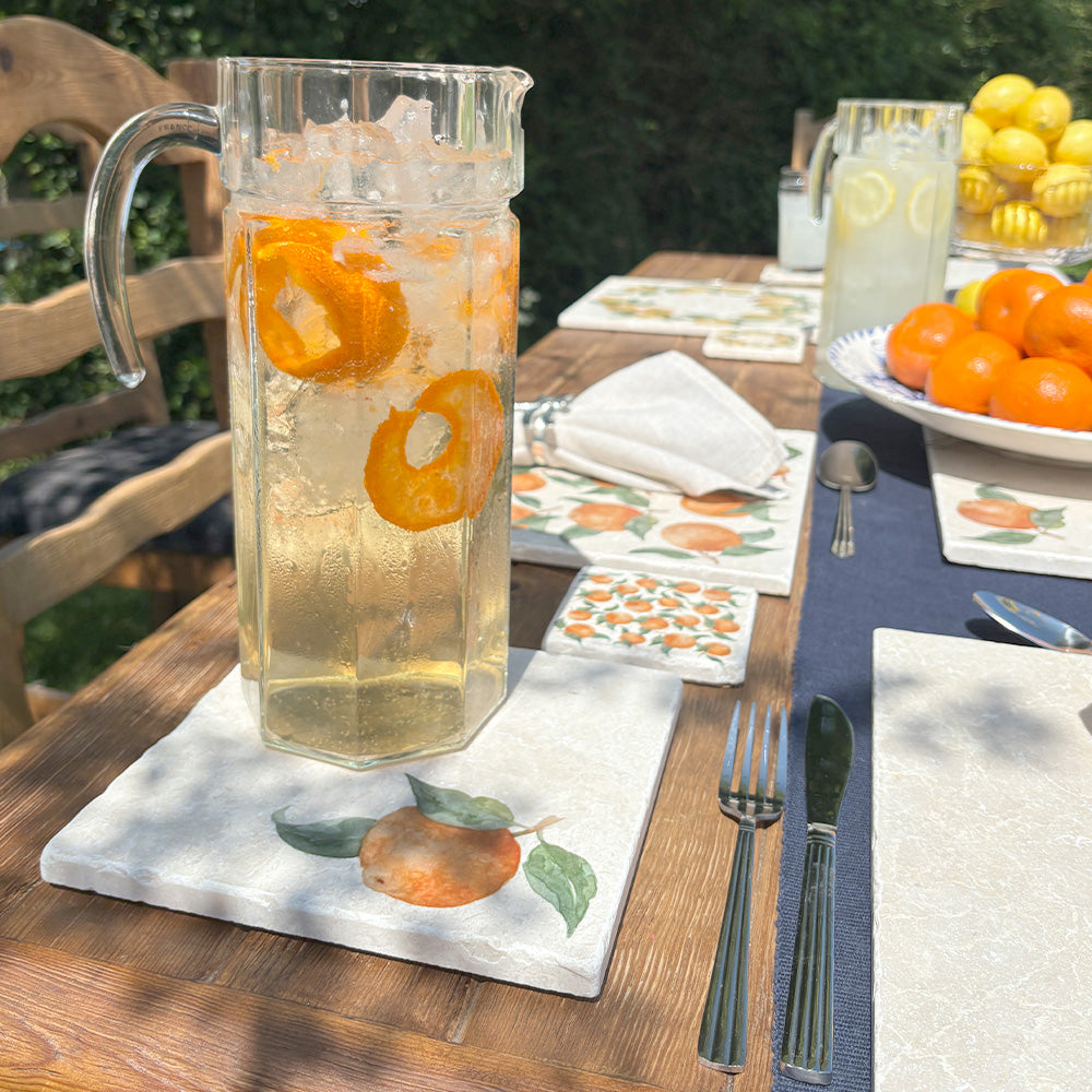 A garden table in the sunshine set with marble placemats, coasters and platters, all featuring a watercolour design of a single orange with leaves.