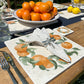 A garden table in the sunshine, the table is set with marble placemats and coasters featuring a rustic orange wreath design.