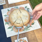 A large marble platter featuring a watercolour orange wreath design.  The platter is being used as a trivet to serve a delicious apple pie.