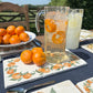 A sunny garden table set with a rectangular marble serving board featuring a watercolour design of oranges and leaves. The sharing platter  has a large jug of orange infused water set on it.