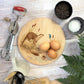 Round wooden chopping board on a kitchen worktop. The beech wood board features a watercolour design of otters and a kingfisher down by the river bank.