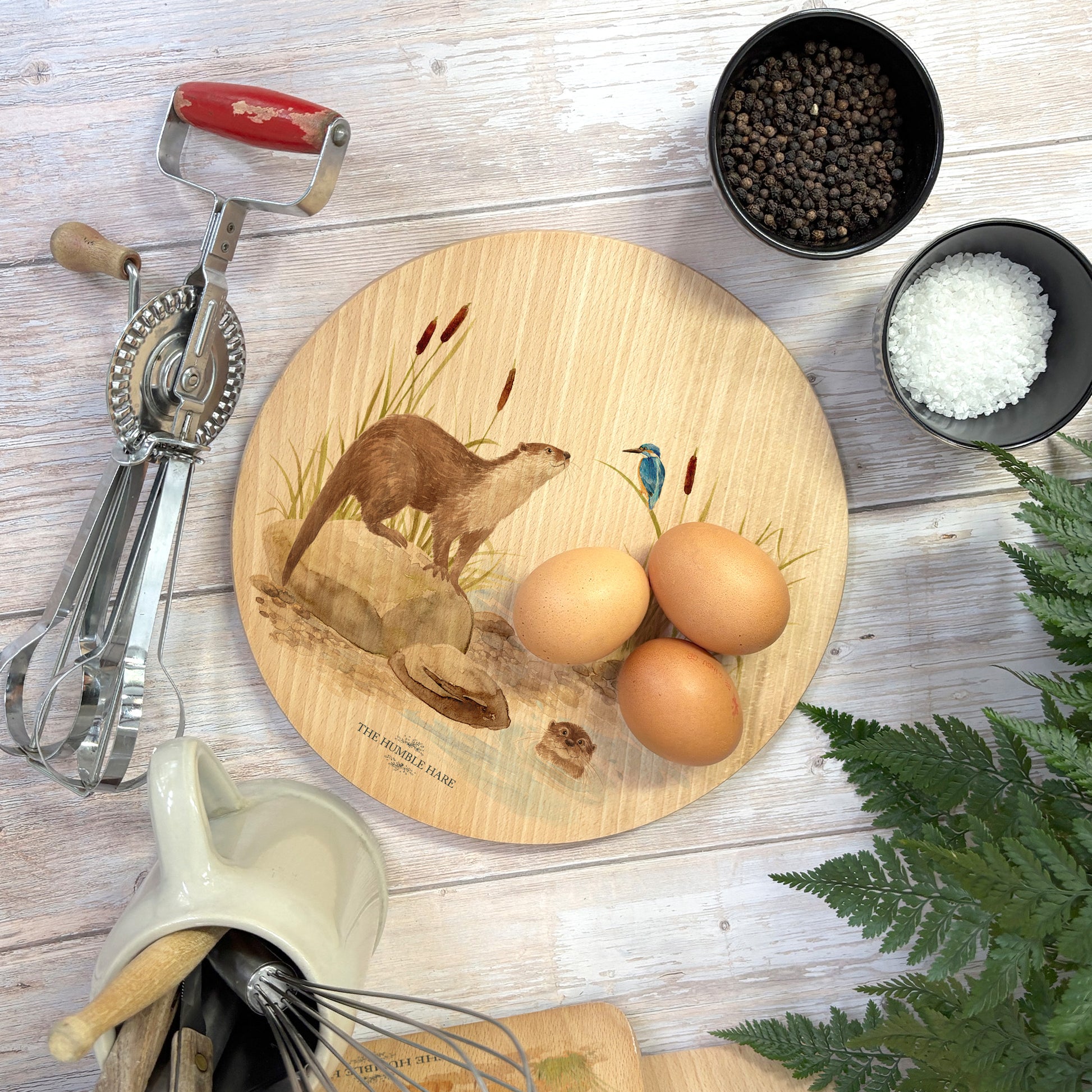 Round wooden chopping board on a kitchen worktop. The beech wood board features a watercolour design of otters and a kingfisher down by the river bank.