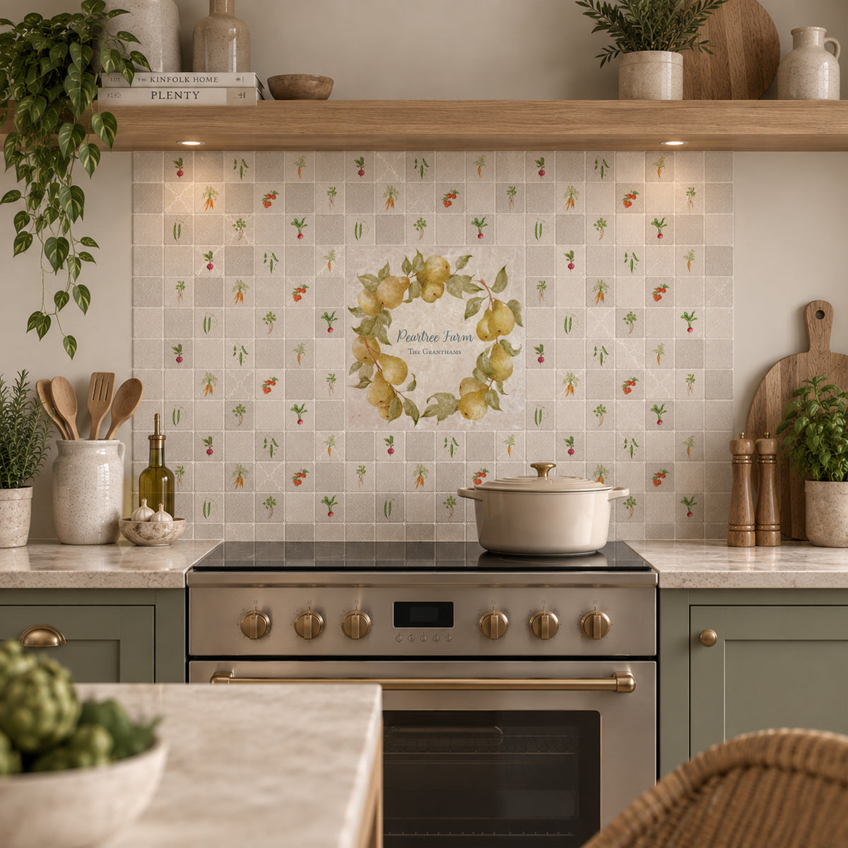 A styled kitchen interior with cream mosaic tile backsplash featuring a central floral wreath graphic and small bird motifs, paired with sage green cabinets, a black stovetop, and warm wood shelving.