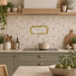 A styled kitchen interior with cream mosaic tile backsplash featuring a central floral wreath graphic and small bird motifs, paired with sage green cabinets, a black stovetop, and warm wood shelving.