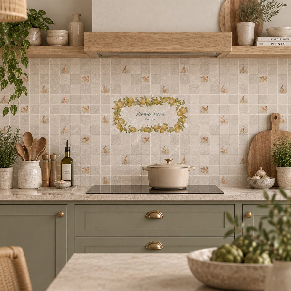A styled kitchen interior with cream mosaic tile backsplash featuring a central floral wreath graphic and small bird motifs, paired with sage green cabinets, a black stovetop, and warm wood shelving.