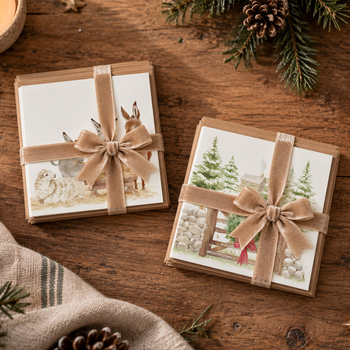 Two packs of square Christmas cards wrapped in a champagne gold ribbon, each pack features a rustic winter watercolour illustration, arranged on a wooden tabletop with pine branches and a candle partially visible.