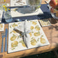 An garden dining table set with marble placemats and coasters featuring a maximalist pattern of golden green pears.