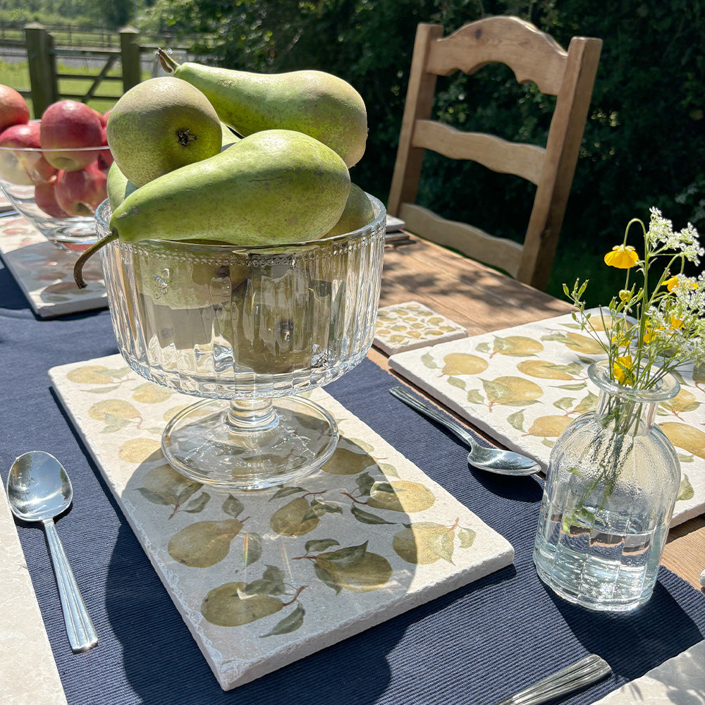 An outdoor table set with elegant bowls of fruit and vases of flowers, placed on multipurpose marble platters featuring a watercolour pattern of golden green pears.