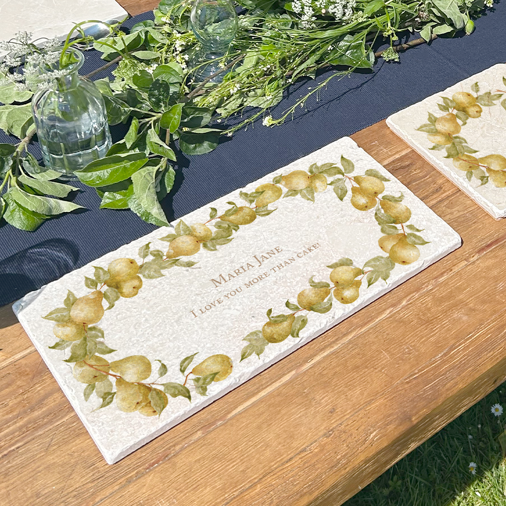 A personalised marble sharing platter featuring a wreath of pears and leaves, with a bespoke message in the centre of the design. The personalised marble platter is placed on a sunny outdoor table.