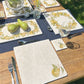 A garden table set for a summer dinner party with handmade marble placemats and coasters featuring a single golden green pear with leaves in a minimalistic design.