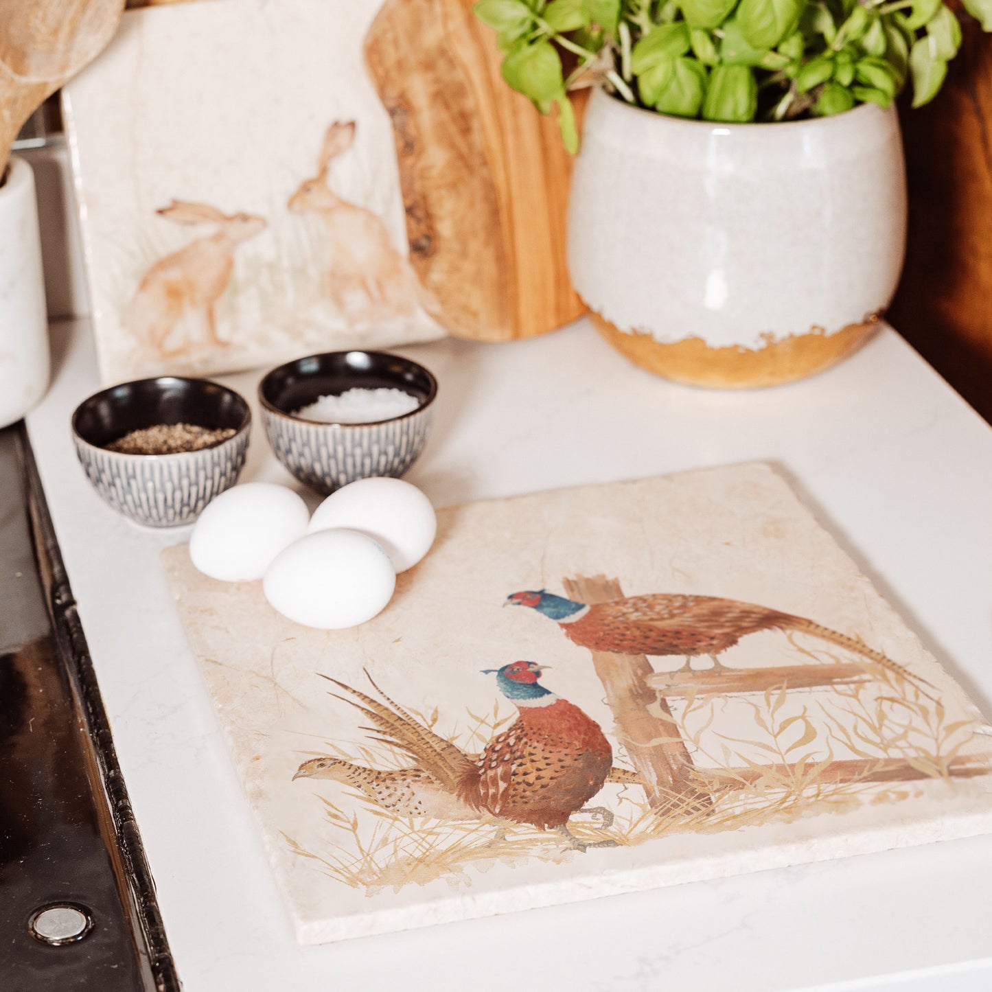 A large square multipurpose marble worktop saver, featuring a watercolour design of three pheasants, two male and one female, standing around a country fence post in golden grass.