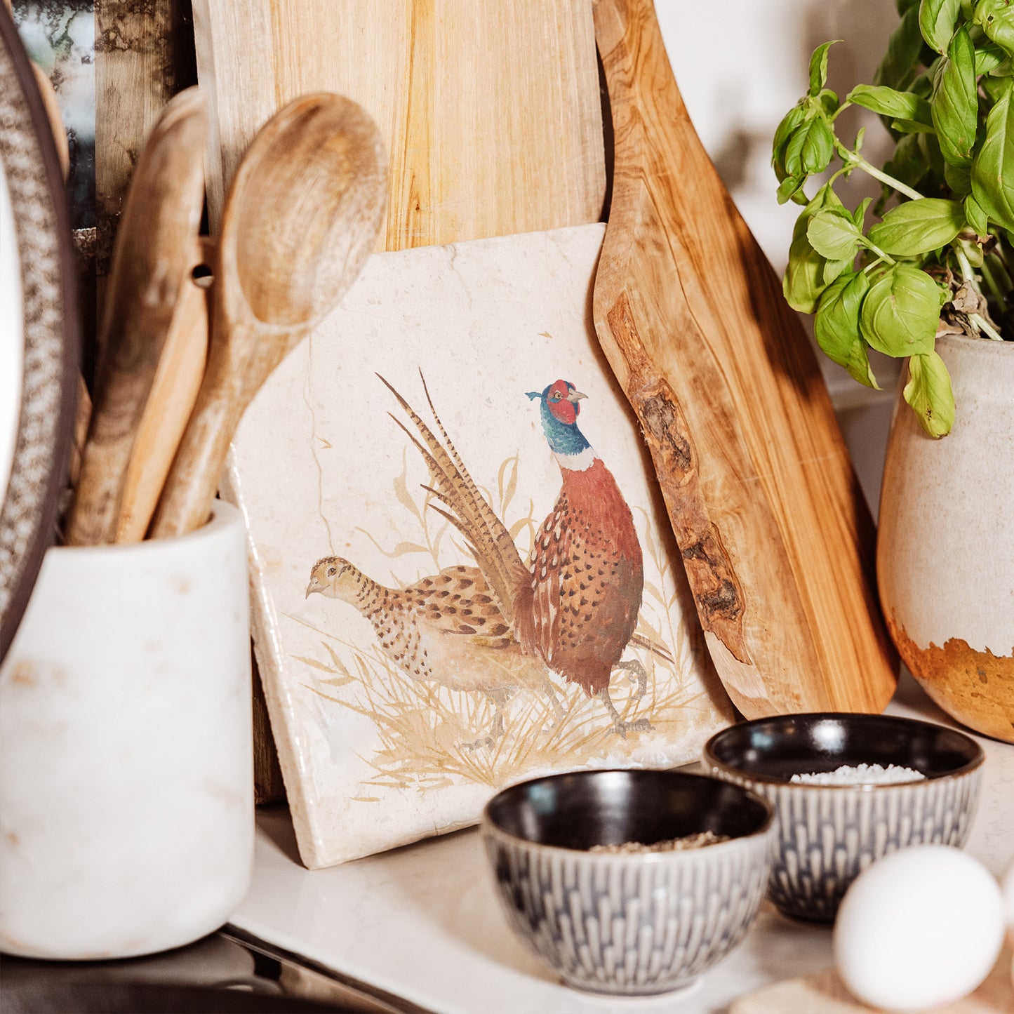 A medium square marble platter featuring a watercolour pheasant design, propped up on a country kitchen worktop.