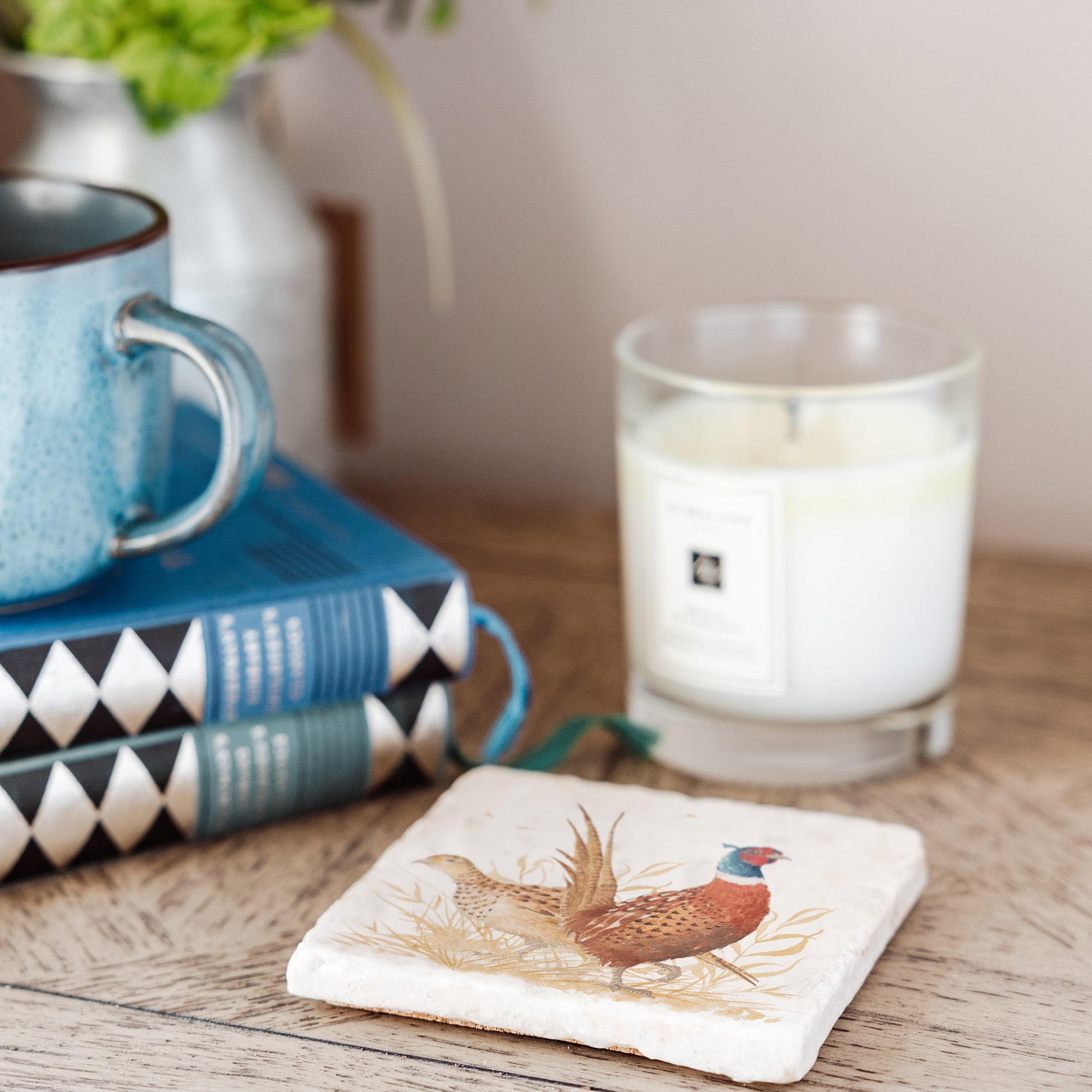 A marble coaster set on a bedside table, the coaster features a watercolour design of a pair of pheasants, one male and one female, in golden grass.