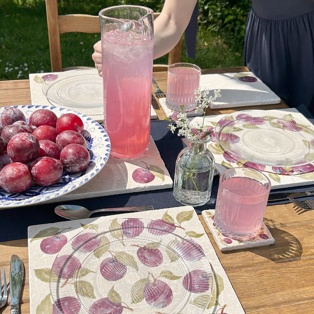 A garden table set with marble placemats and coasters and vibrant cocktails, each marble platter featuring a maximalist plum pattern.