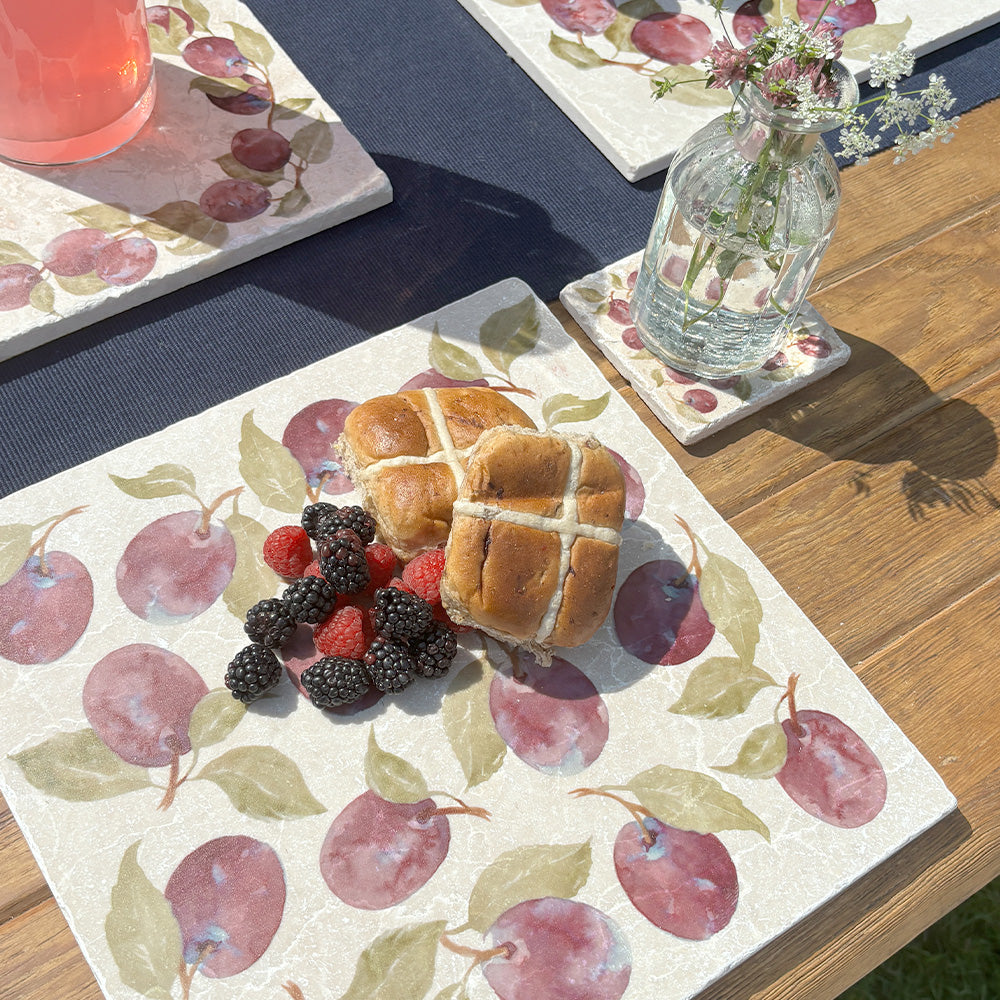 A garden dinner table set with cream marble placemats and coasters featuring a maximalist purple plum pattern.