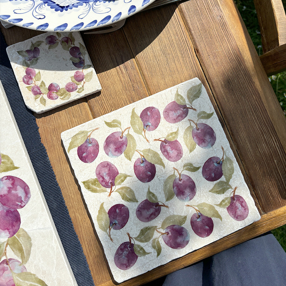 A garden table in the sunshine set with marble placemats, coasters and platters, all featuring a watercolour plum design. 